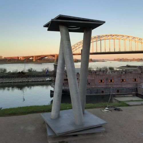 Skulptur am Ufer mit Fluss und Brücke im Hintergrund bei Sonnenuntergang