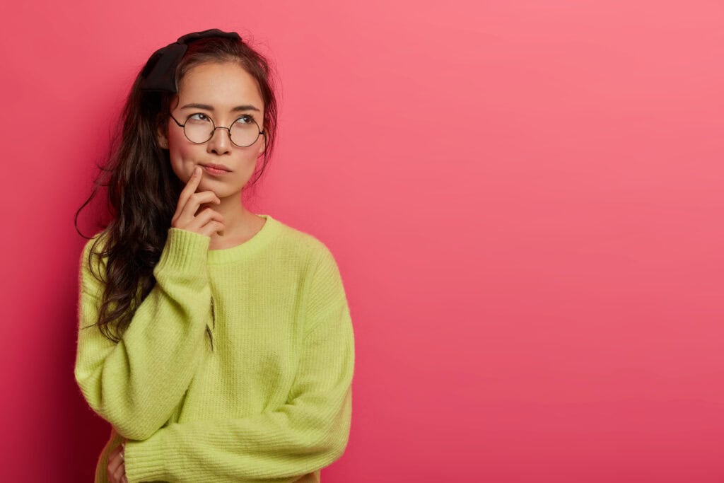 Eine nachdenkliche junge Frau mit Brille und grünem Pullover vor einem pinken Hintergrund.