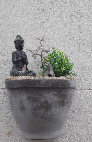 Kleine Buddha-Statue in Meditationshaltung, platziert in einem rustikalen Blumentopf mit einem Bonsai-Bäumchen und grünen Pflanzen, vor einer grauen Wand.