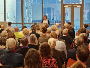 Ein Foto der Zen-Lehrerin am Rednerpult während ihres Vortrags. Sie hat lockiges, braunes Haar und trägt eine schwarze Jacke über einem weißen Oberteil. Der Hintergrund zeigt eine große Fensterfront mit Birkenbäumen und das Publikum im Vordergrund.